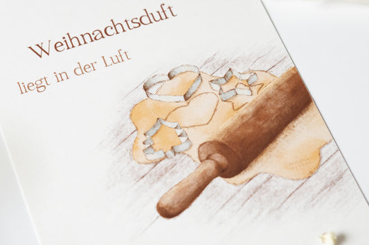 Postkarte „Weihnachtsduft liegt in der Luft“ mit Illustration von Plätzchenteig, Ausstechformen und Nudelholz