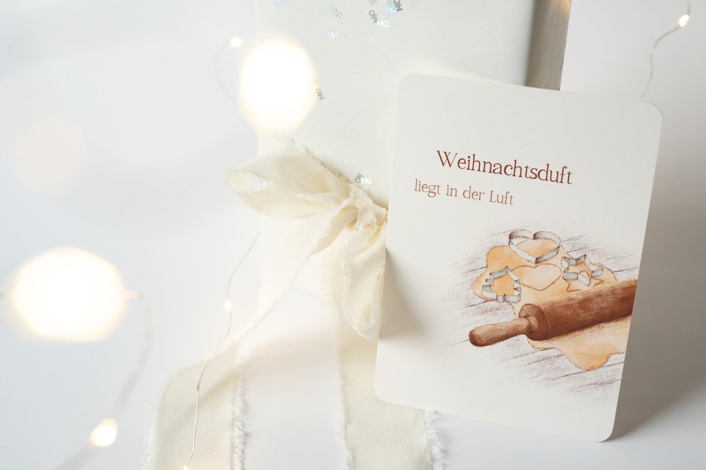 Postkarte „Weihnachtsduft liegt in der Luft“ mit Illustration von Plätzchenteig, Ausstechformen und Nudelholz