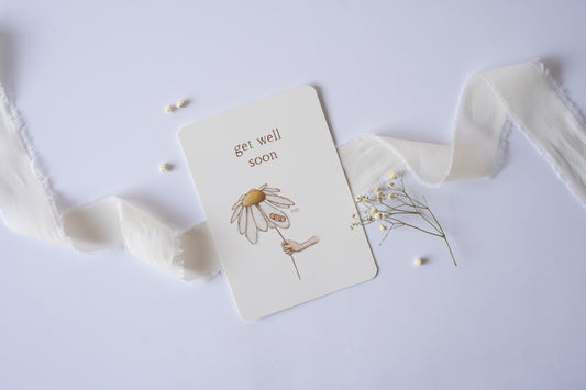 Postkarte mit zarter handgezeichneter Blume und Pflaster, darüber der Spruch „Get Well Soon“ – liebevolle Gute-Besserungskarte auf Naturpapier.