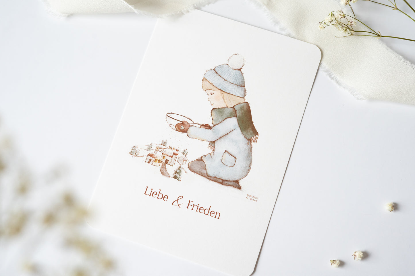 Postkarte mit winterlicher Illustration eines Kindes vor einem Dorf und dem Text „Liebe & Frieden“, Weihnachtskarte A6 von FranziGruDesign