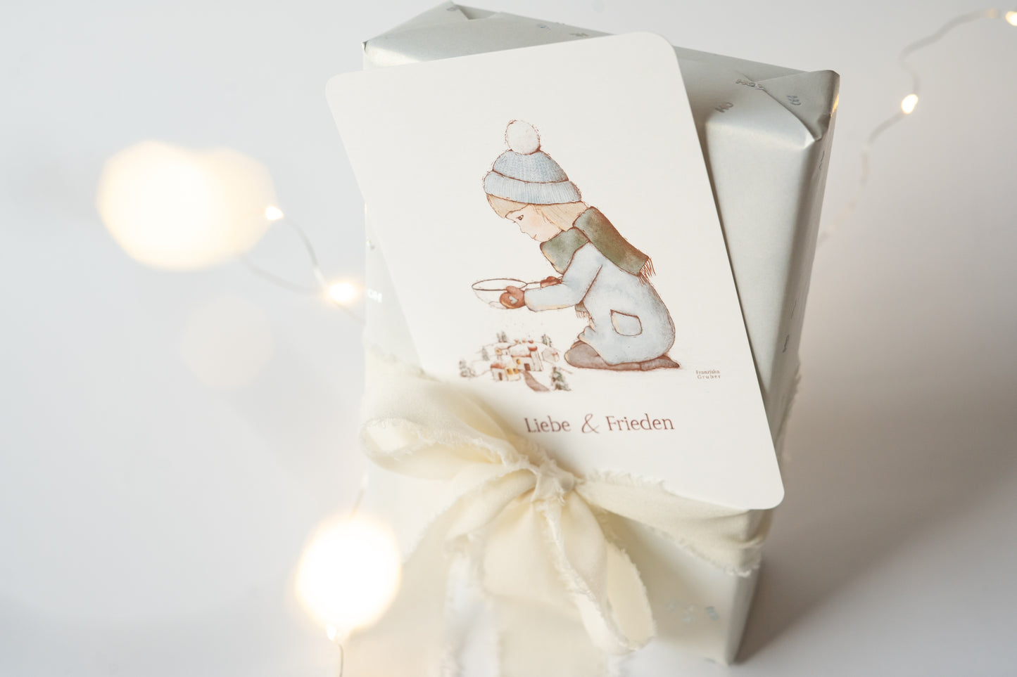 Postkarte mit winterlicher Illustration eines Kindes vor einem Dorf und dem Text „Liebe & Frieden“, Weihnachtskarte A6 von FranziGruDesign