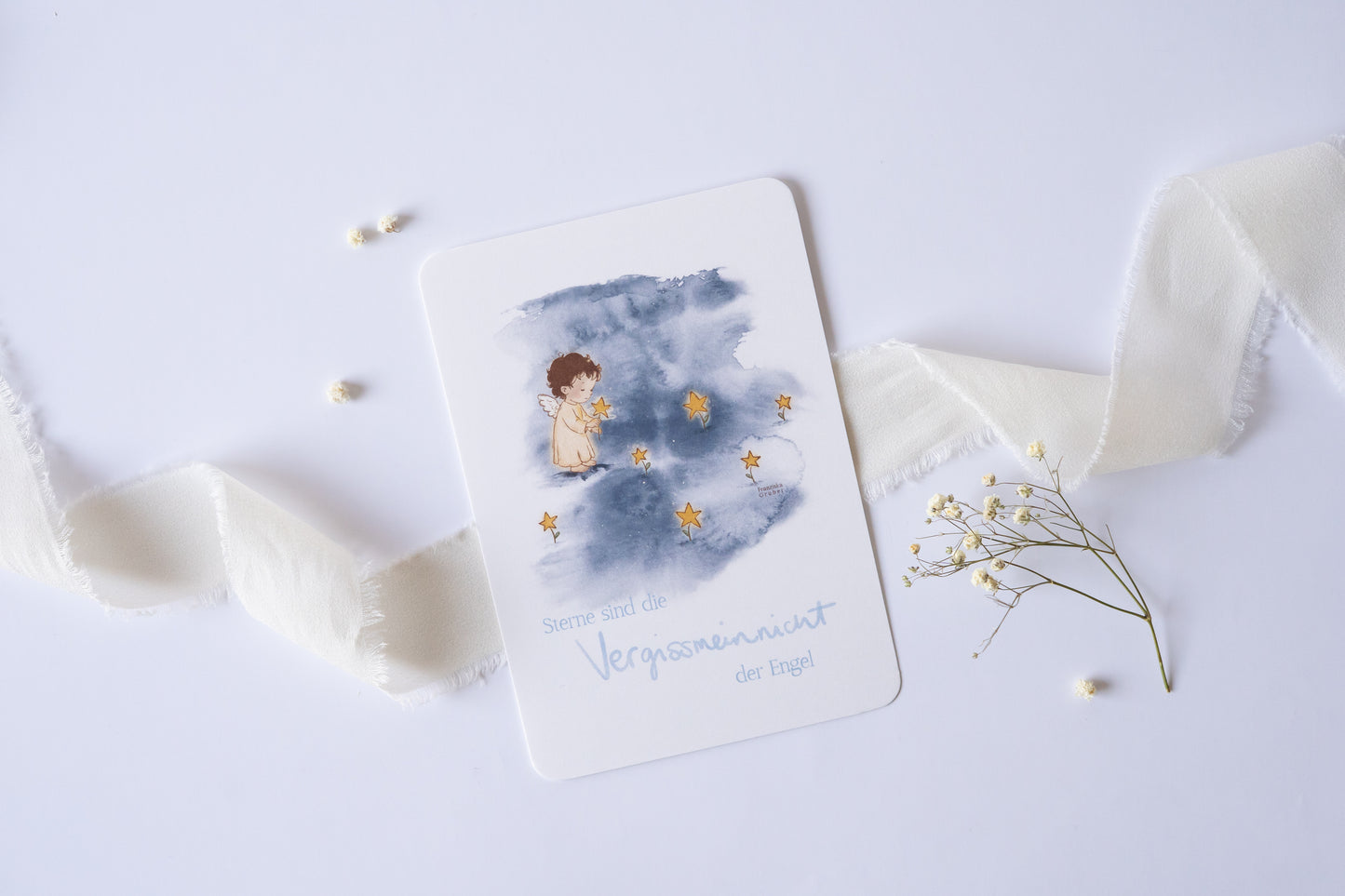 Postkarte mit Engel und goldenen Sternen vor blauem Aquarellhintergrund und dem Spruch „Sterne sind die Vergissmeinnicht der Engel“ – Trauer- und Gedenkkarte.