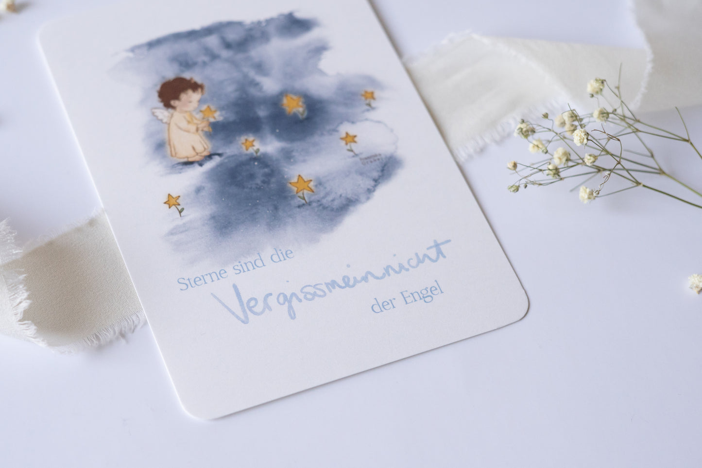 Postkarte mit Engel und goldenen Sternen vor blauem Aquarellhintergrund und dem Spruch „Sterne sind die Vergissmeinnicht der Engel“ – Trauer- und Gedenkkarte.