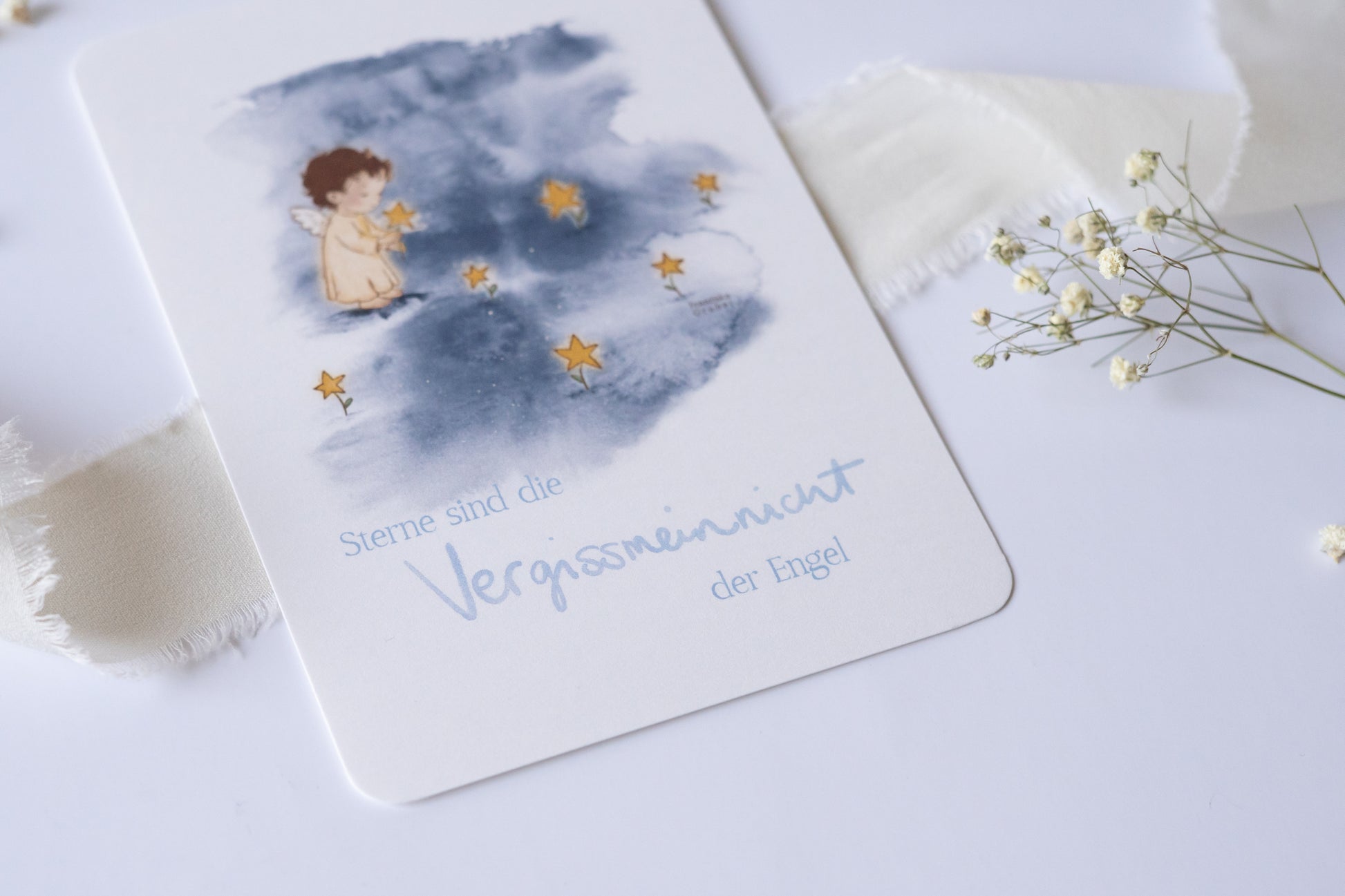 Postkarte mit Engel und goldenen Sternen vor blauem Aquarellhintergrund und dem Spruch „Sterne sind die Vergissmeinnicht der Engel“ – Trauer- und Gedenkkarte.