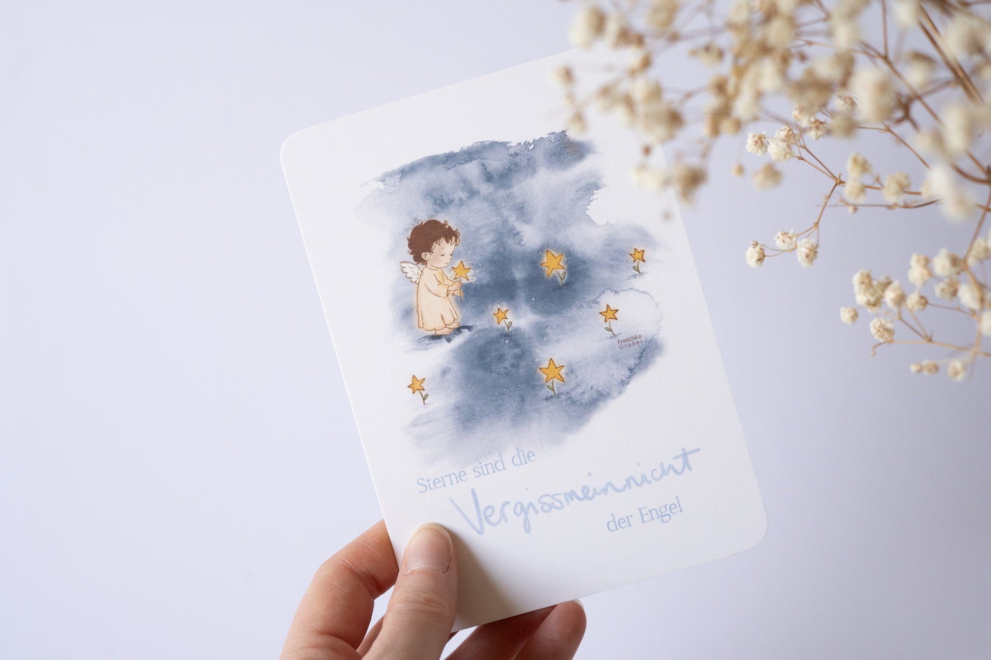 Postkarte mit Engel und goldenen Sternen vor blauem Aquarellhintergrund und dem Spruch „Sterne sind die Vergissmeinnicht der Engel“ – Trauer- und Gedenkkarte.