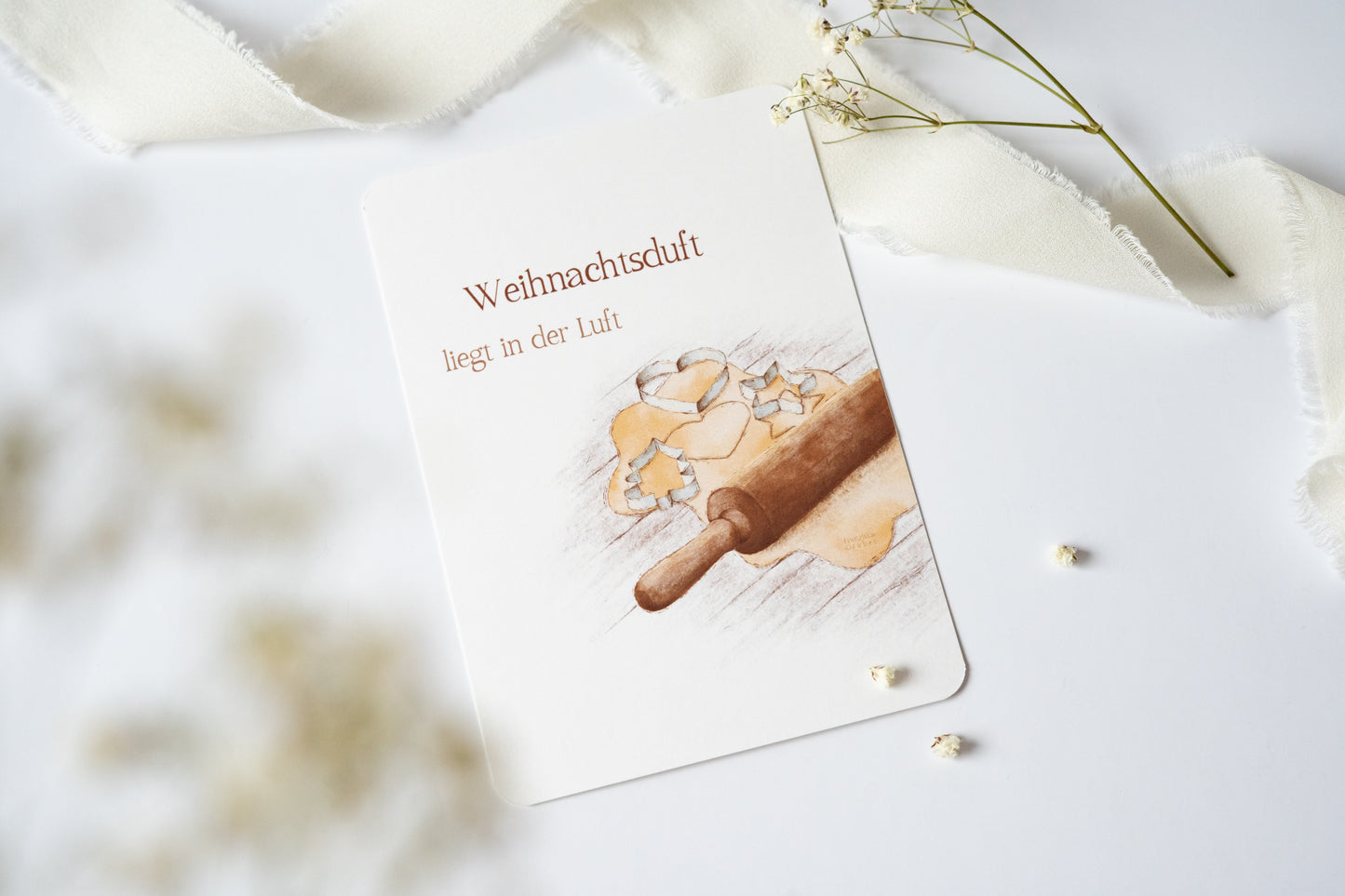 Postkarte „Weihnachtsduft liegt in der Luft“ mit Illustration von Plätzchenteig, Ausstechformen und Nudelholz