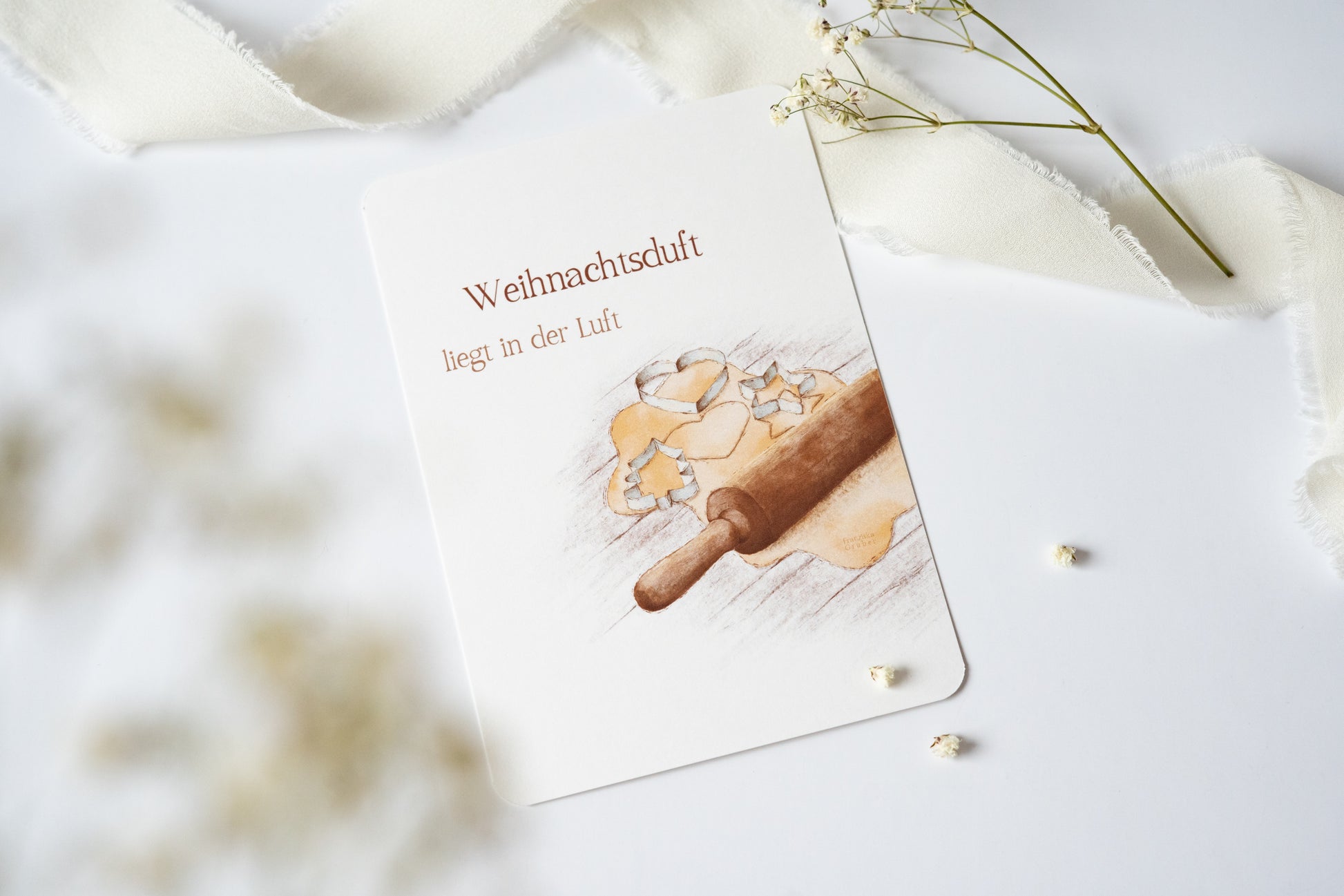 Postkarte „Weihnachtsduft liegt in der Luft“ mit Illustration von Plätzchenteig, Ausstechformen und Nudelholz