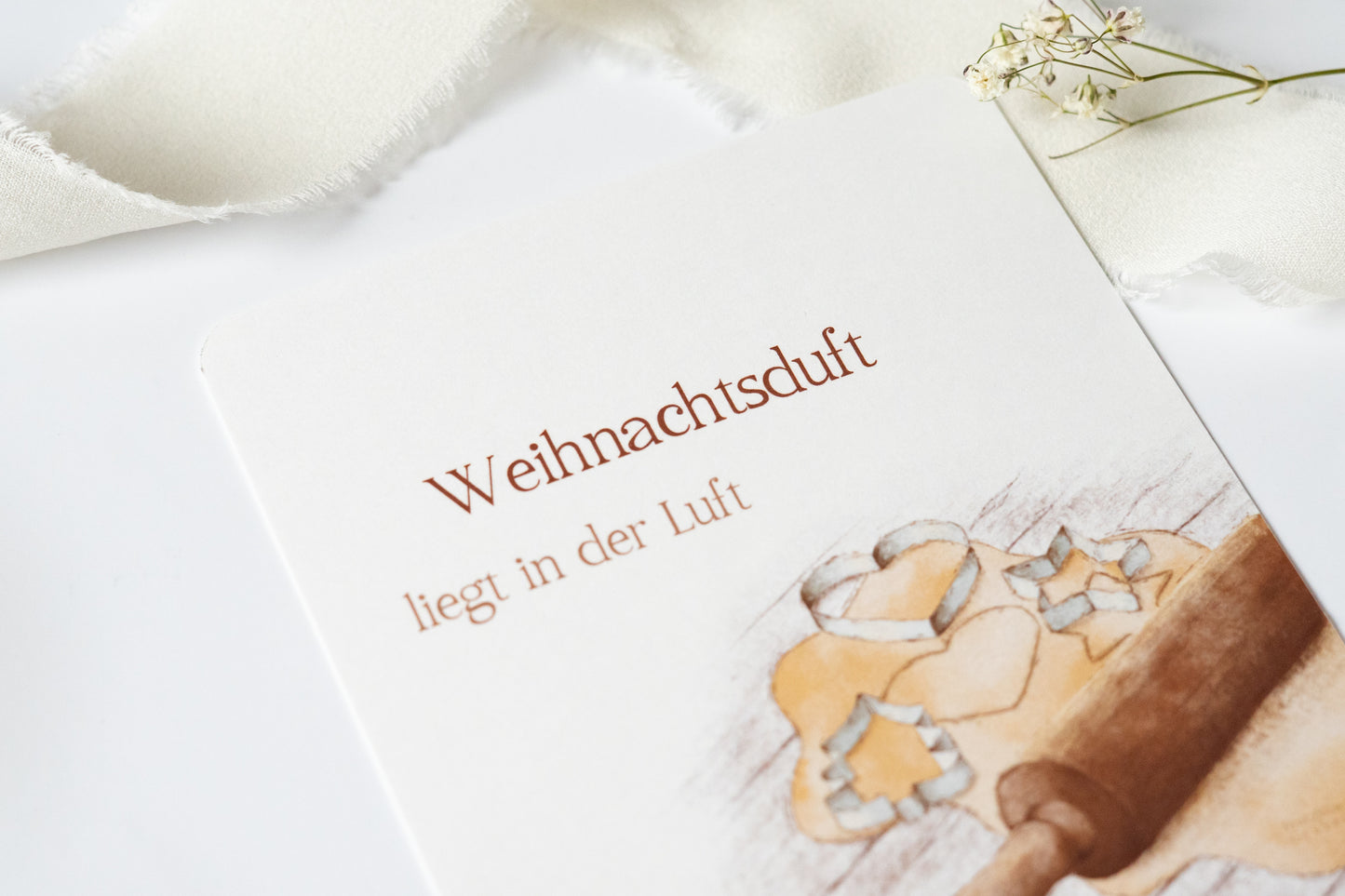 Postkarte „Weihnachtsduft liegt in der Luft“ mit Illustration von Plätzchenteig, Ausstechformen und Nudelholz