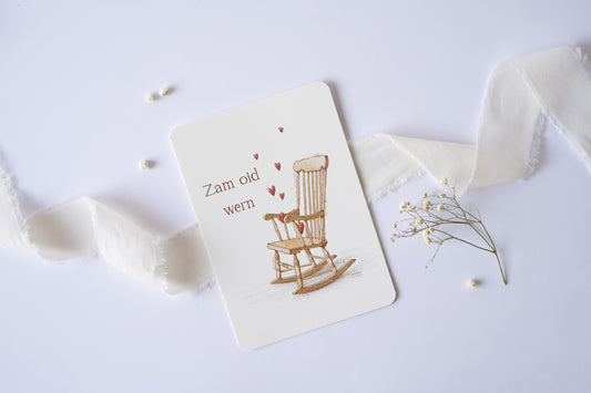 Postkarte „Zam oid wern“ im bayerischen Dialekt mit handgezeichneter Schaukelstuhl-Illustration und roten Herzen – ideal für Hochzeit oder Jahrestag.